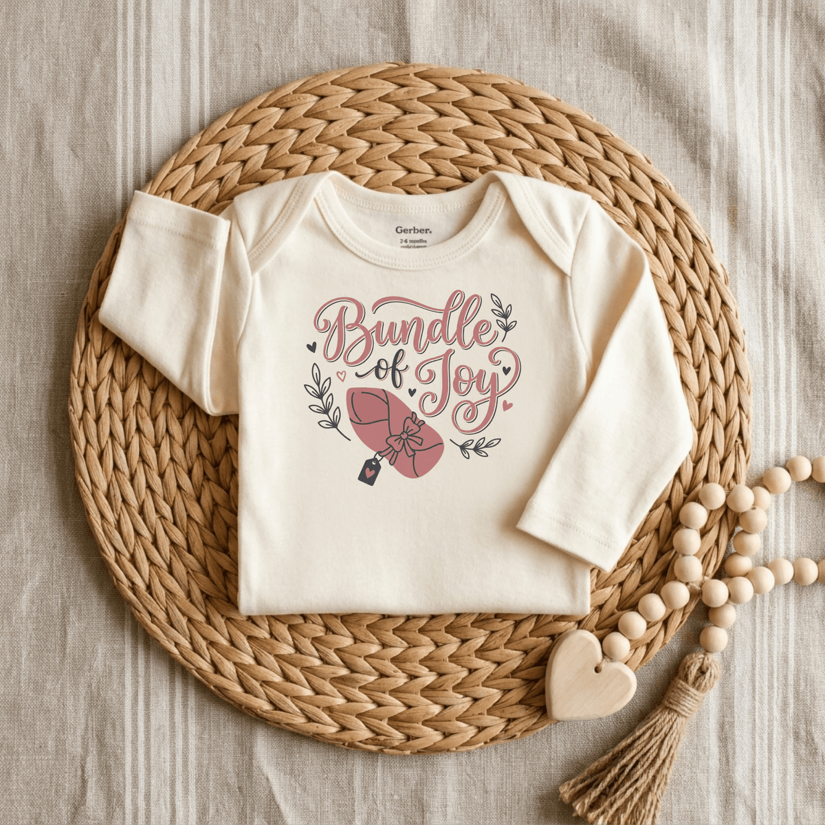 The Toodleroos Bundle of Joy Baby Girl Onesie – Gift-Wrapped Bodysuit features a cream design with “Bundle of Joy” and a pink bundle illustration, arranged on a woven mat beside a wooden bead and heart garland. This cozy Gerber bodysuit is the perfect baby shower gift, presented against a neutral, inviting background.
