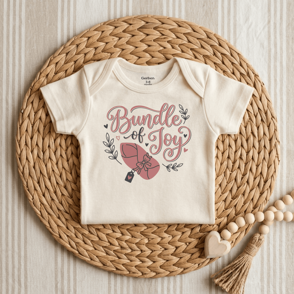 A Toodleroos Bundle of Joy Baby Girl Onesie – Gift-Wrapped Bodysuit (size 3-6 months) in a soft off-white hue is artfully placed on a circular woven placemat. Ideal as a thoughtful baby shower gift, this Gerber bodysuit showcases the phrase “Bundle of Joy” in elegant script, accented with charming illustrations of a swaddled baby, delicate leaves, and tiny hearts in pink and grey.