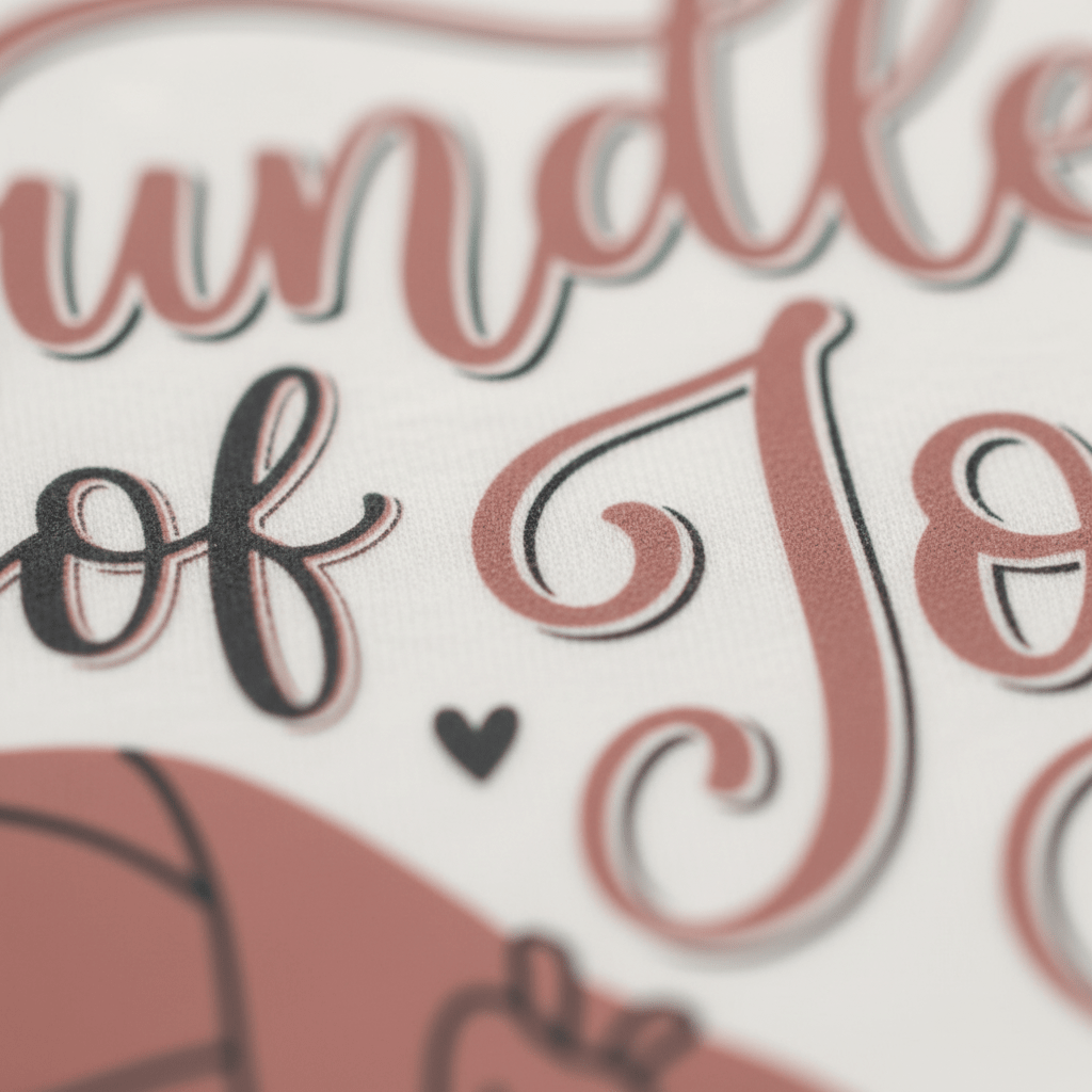 Close-up of stylized text on fabric reading bundle of joy in cursive font, with a small heart symbol and part of a graphic visible at the bottom—perfect as the Toodleroos Bundle of Joy Baby Girl Onesie – a gift-wrapped bodysuit ideal for baby showers or welcoming your little one.