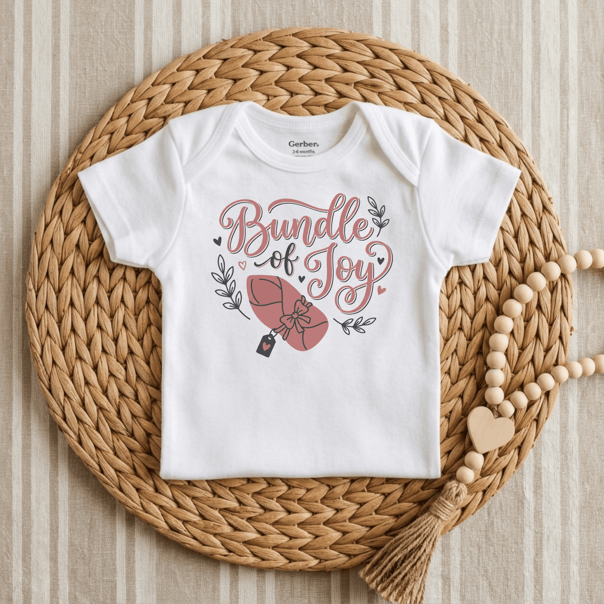 A Toodleroos Bundle of Joy Baby Girl Onesie – Gift-Wrapped Bodysuit in crisp white, featuring a charming wrapped baby illustration, rests on a woven placemat beside a wooden-beaded garland with heart and tassel accents—making it the perfect adorable baby shower gift.