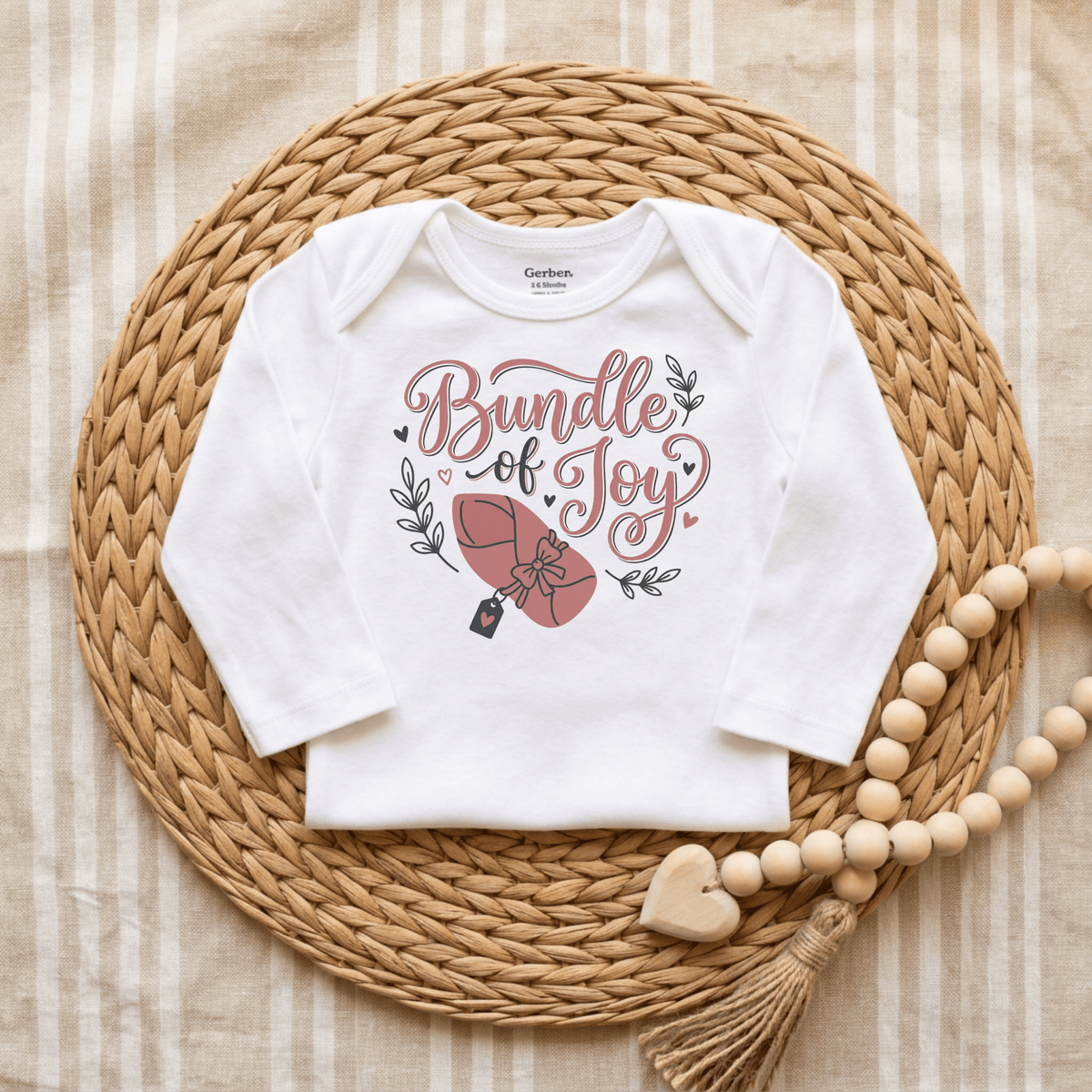 A Toodleroos Bundle of Joy Baby Girl Onesie – Gift-Wrapped Bodysuit, adorned with a “Bundle of Joy” and umbrella design, is displayed on a woven placemat with wooden bead decor against a striped fabric background—making it an adorable choice for baby shower gifting.