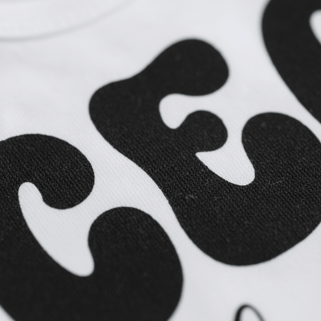 Close-up of bold, black, curvy lettering printed on white fabric—only parts of the playful “CEO of the House” text are visible, showcasing the textured material and striking contrast. This detail highlights the fun, gender-neutral style of Toodleroos’ CEO of the House Tee—a funny shirt for babies, toddlers, and young trendsetters alike.