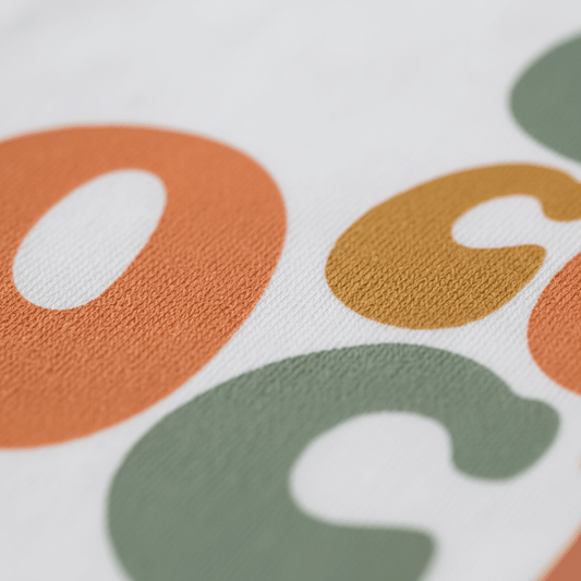 Closeup of colorful, large, rounded letters O, C, and C in orange, mustard yellow, and green printed on textured white fabric. These playful letters are the highlight of the Toodleroos Cool Cousins Club Toddler Shirt – Retro Family Tee—an ideal pick for family reunions or toddler outings.
