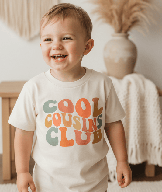 A happy toddler wears the Toodleroos Cool Cousins Club Toddler Shirt – Retro Family Tee in a soft cream shade with vibrant lettering, standing indoors against cozy neutral decor—an ideal matching cousin shirt for your next family reunion.