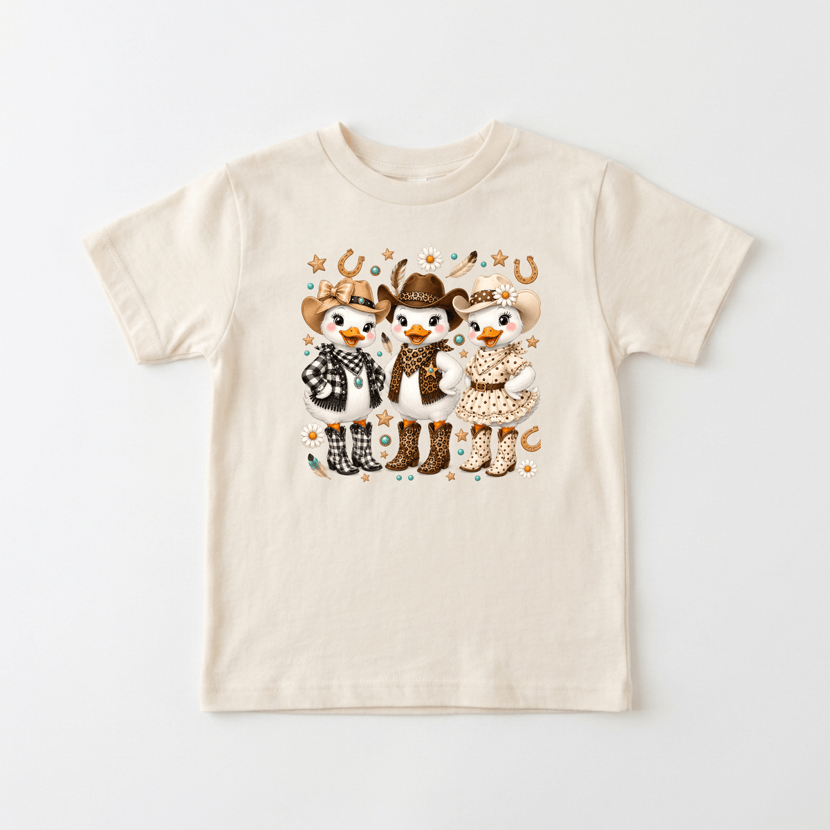 The Toodleroos Cowgirl Duck Trio Kids Shirt – Country Girls Tee is a beige t-shirt featuring three cartoon animals—dressed as cowboys and cowgirls in hats, boots, and western outfits—surrounded by horseshoes and stars on a plain background. Perfect for fans of country kids shirts or anyone who loves a fun western twist on a Cowgirl duck trio shirt.