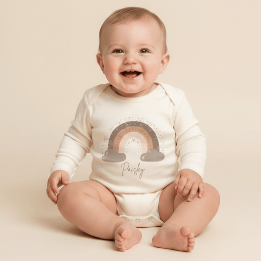 A smiling baby sits on a light background, wearing the Toodleroos Custom Boho Rainbow Newborn Onesie – a cream-colored bodysuit featuring a charming rainbow and clouds design. The name Paisley is printed below the rainbow, making this personalized name bodysuit an ideal custom baby shower gift.