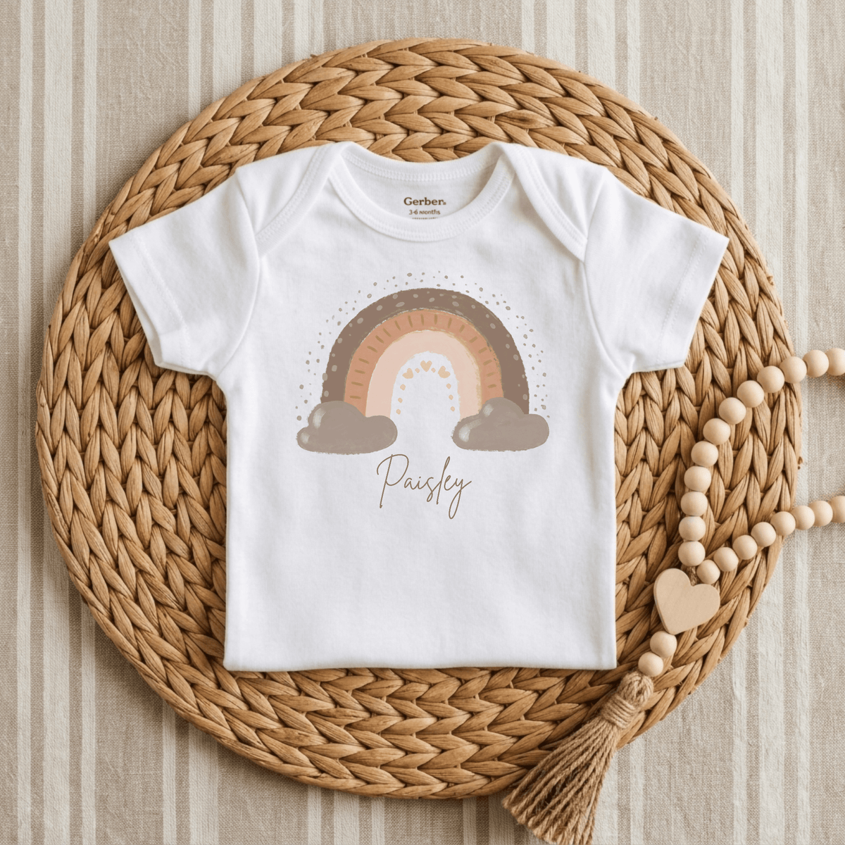 A Custom Boho Rainbow Newborn Onesie – Personalized Name Bodysuit by Toodleroos in white, sized 3–6 months, is displayed on a round woven mat. This charming baby bodysuit features a stylish brown rainbow and cloud graphic with the name Paisley printed below. Wooden beads with a heart charm rest to the right on a striped fabric background, making it an ideal custom baby shower gift.
