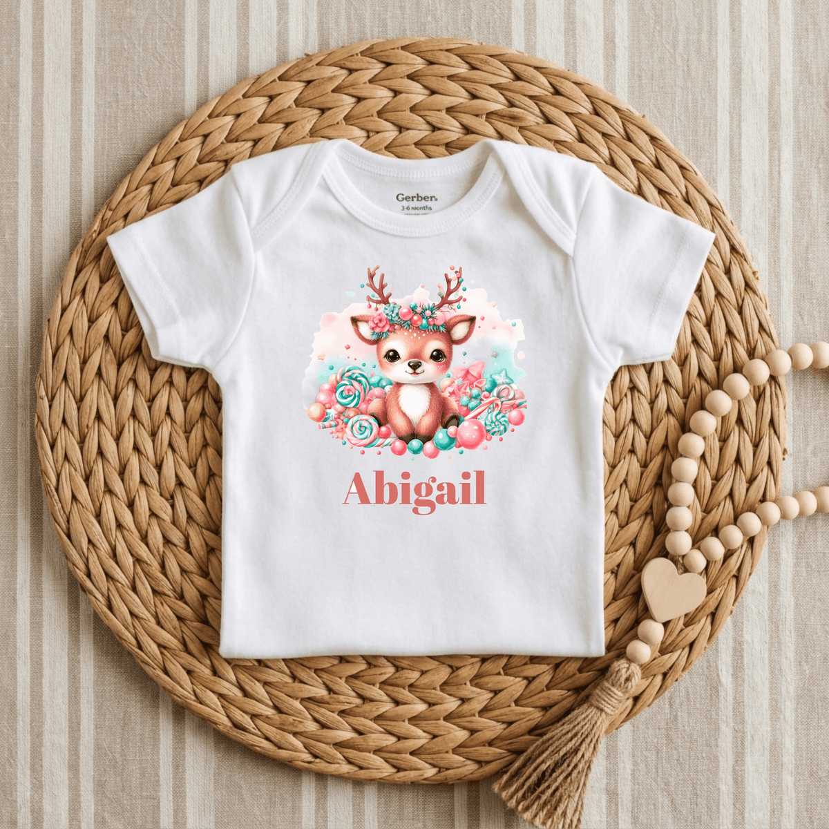 The Toodleroos Custom Candy Deer Baby Onesie – Personalized Name Bodysuit showcases a charming deer illustration surrounded by lovely flowers, with the name Abigail printed on it. Displayed on a round woven placemat beside a beaded wooden garland with a heart charm, this adorable bodysuit makes an ideal custom baby shower gift.