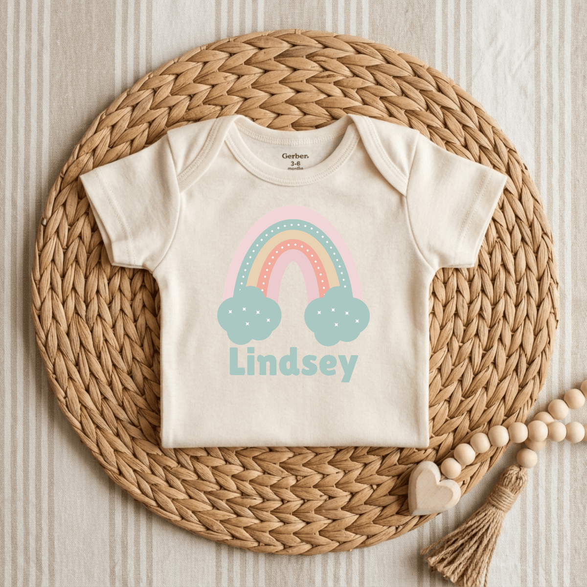 A cream-colored Toodleroos Custom Pastel Rainbow Infant Onesie – Personalized Name Bodysuit in size 3-6 months is centered on a woven rattan placemat. The soft, custom baby bodysuit showcases a cheerful pastel rainbow design with blue clouds and “Lindsey” printed in blue letters beneath the graphic. Nearby, wood beads and a heart accentuate the setup, making this charming piece a thoughtful choice for a baby shower gift.