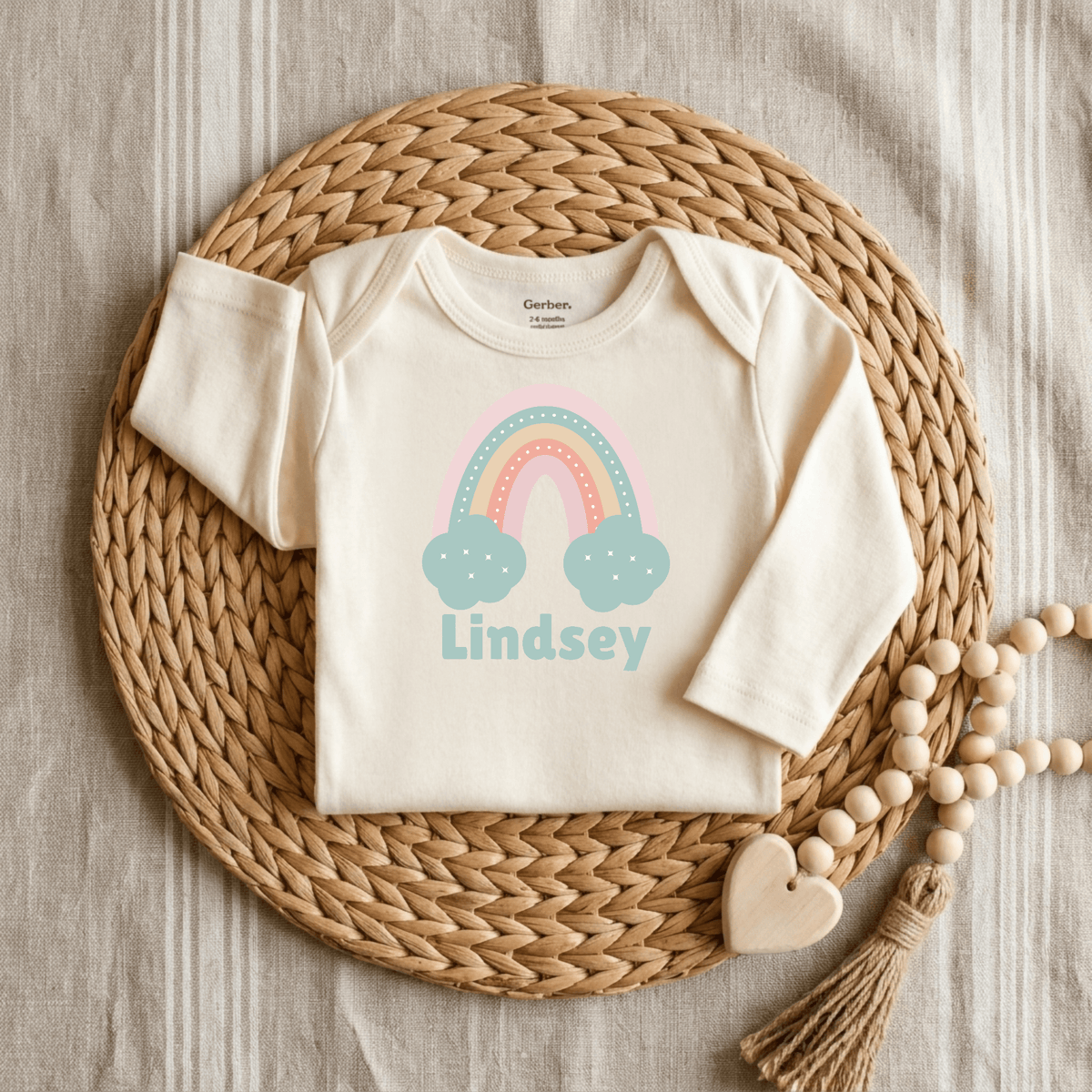 Introducing the Toodleroos Custom Pastel Rainbow Infant Onesie – Personalized Name Bodysuit: A cream-colored baby onesie featuring charming pastel rainbow and blue clouds artwork, customized with the name Lindsey across the front. Beautifully displayed on a woven mat with a wooden beaded garland and heart accent, this personalized bodysuit makes an ideal baby shower gift.