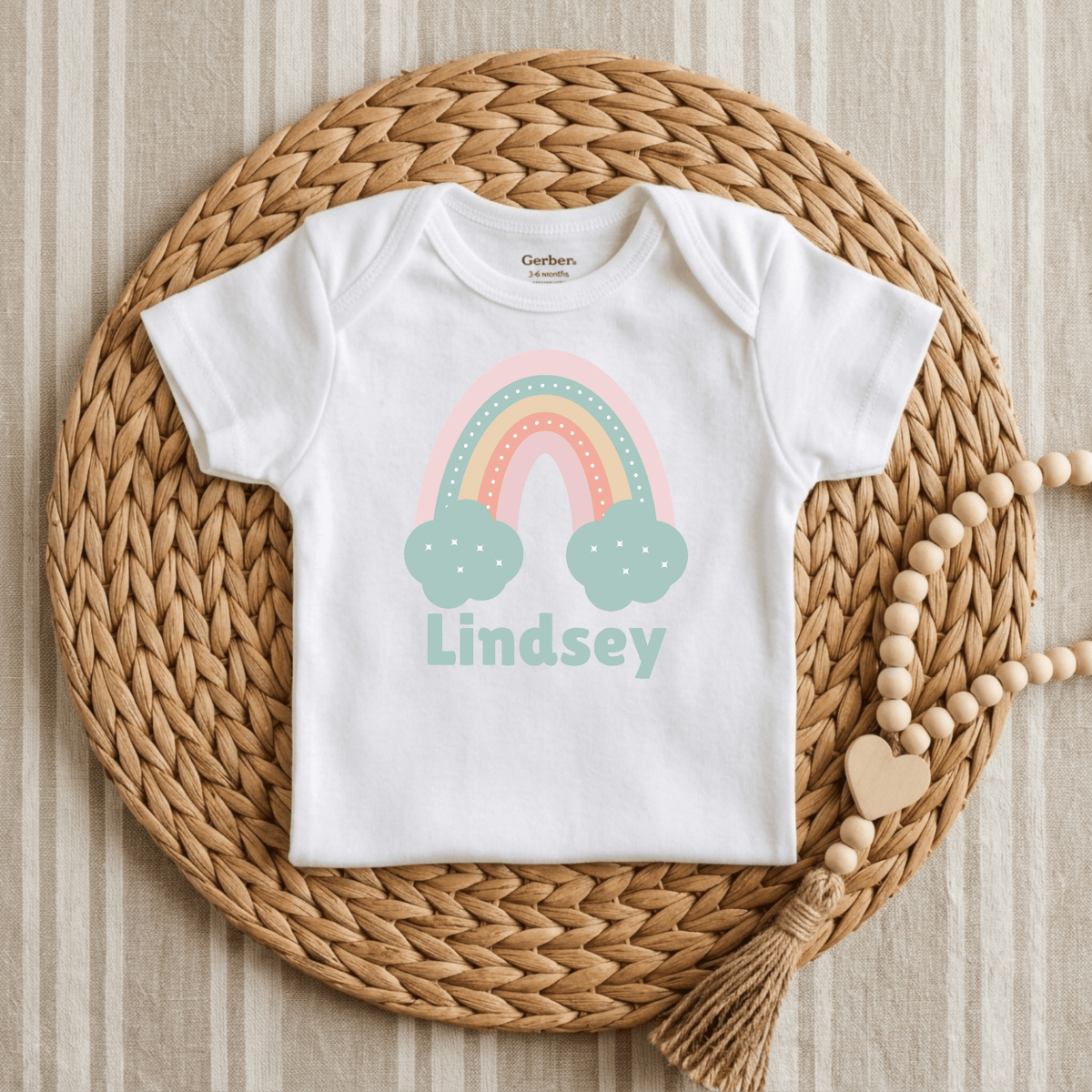 The Toodleroos Custom Pastel Rainbow Infant Onesie – Personalized Name Bodysuit is shown on a woven placemat, adorned with a beaded tassel garland. This adorable baby shower gift features a pastel rainbow and clouds design, customized with the name Lindsey printed on it.