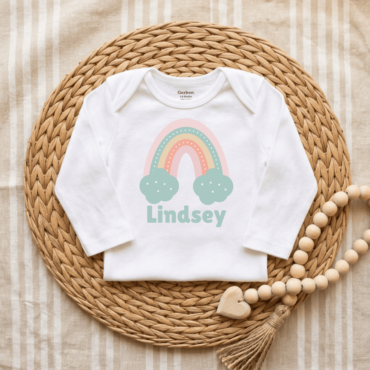 Introduce a touch of charm with the Custom Pastel Rainbow Infant Onesie – Personalized Name Bodysuit by Toodleroos. This white custom baby bodysuit features a soft pastel rainbow and clouds design, personalized with the name “Lindsey” underneath. Beautifully displayed on a woven mat alongside a wooden bead garland and set against a striped fabric background, it’s an ideal choice for a thoughtful baby shower gift or unique personalized onesie.
