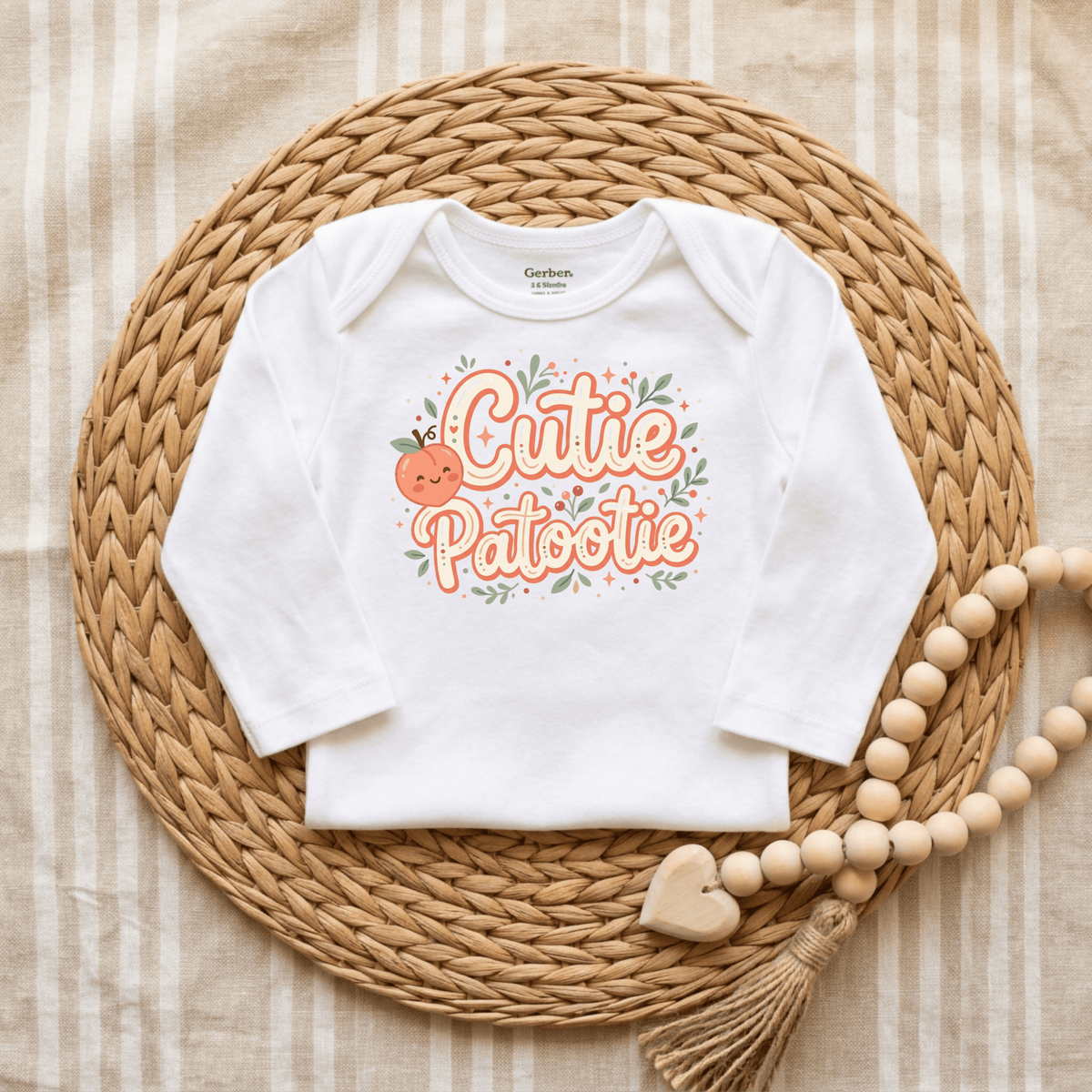 A white, gender-neutral Cutie Patootie Baby Onesie – Sweet Peach Bodysuit by Toodleroos, featuring “Cutie Patootie” and a sweet peach design, is displayed on a round woven mat beside a wooden bead garland with a heart and tassel. The background is a striped cloth, making this adorable Toodleroos onesie the perfect baby shower gift.