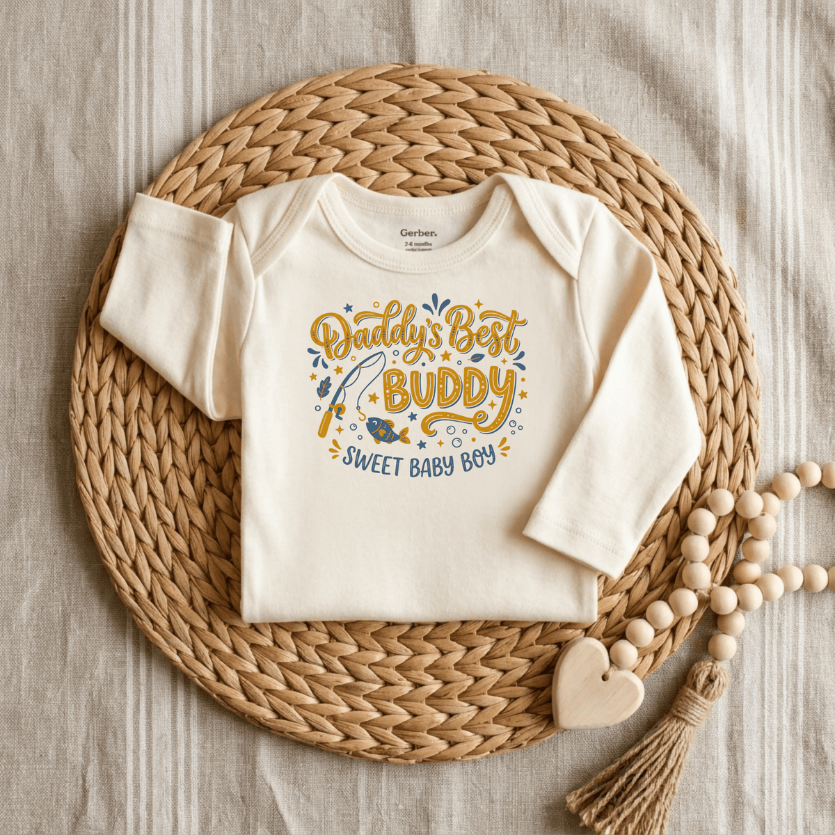 A cream-colored Daddy’s Best Buddy Baby Onesie – Fishing Buddy Bodysuit by Toodleroos is displayed on a woven mat, featuring yellow and blue text that reads “Daddys Best Buddy, Sweet Baby Boy.” Perfect as a Father’s Day baby gift, the onesie is pictured next to a beaded toy with a wooden heart.