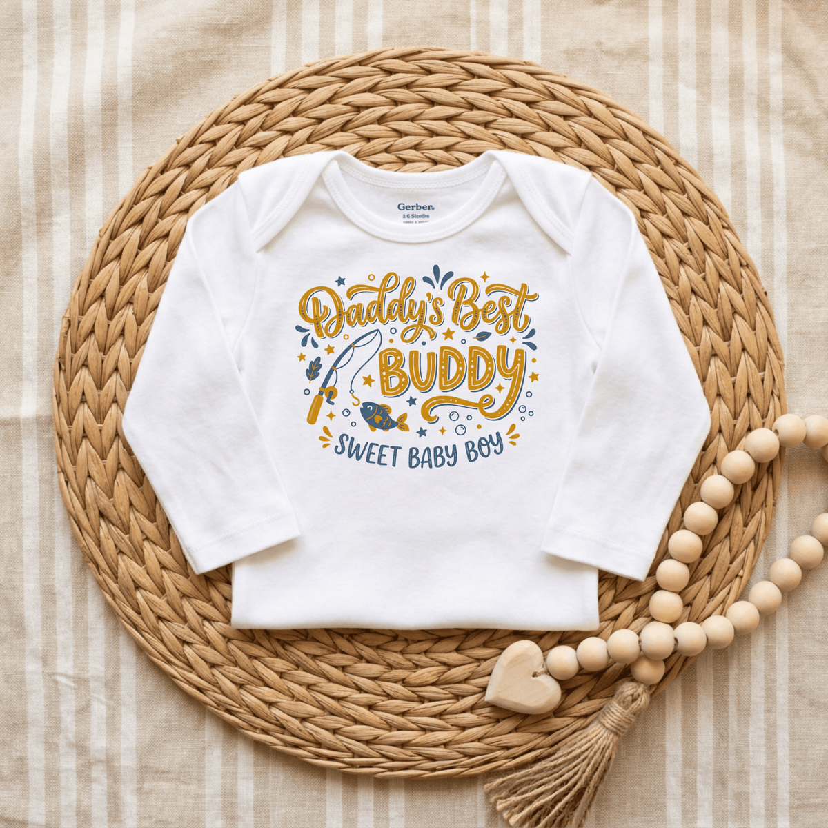 A white Toodleroos Daddy’s Best Buddy Baby Onesie – Fishing Buddy Bodysuit featuring the text “Daddy’s Best Buddy, Sweet Baby Boy” in blue and yellow lettering, perfect as a Father’s Day baby gift. The onesie is displayed on a woven mat with a beaded garland and heart-shaped decoration beside it.