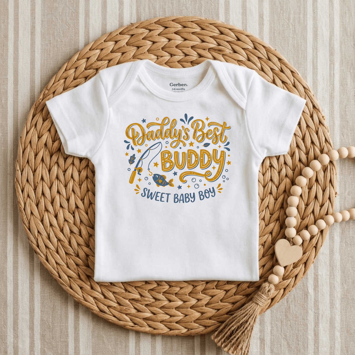 A white Daddy’s Best Buddy Baby Onesie – Fishing Buddy Bodysuit by Toodleroos, perfect as a Father’s Day baby gift, showcases a fishing-themed graphic with the phrases “Daddy’s Best BUDDY” and “SWEET BABY BOY” in blue and mustard yellow script. The adorable onesie is displayed centered on a woven rattan placemat atop a striped fabric background, accented with wooden beads.