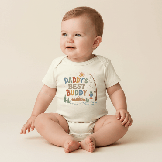 A smiling baby sits on the floor wearing the Toodleroos Baby Boy Fishing Onesie – Daddy’s Best Buddy Short-Sleeve Bodysuit, featuring a charming fishing-themed design and the phrase “Daddy’s Best Buddy” printed on the front.