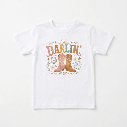 Dress your little one for western adventures with the Toddler Cowgirl Shirt – Darlin’ Western Cowboy Boots Short-Sleeve Tee from Toodleroos. This adorable tee features “Darlin’” in colorful letters above a pair of pink and brown cowboy boots, surrounded by pastel flowers, stars, a horseshoe, and leaves—perfect for any toddler cowgirl!.