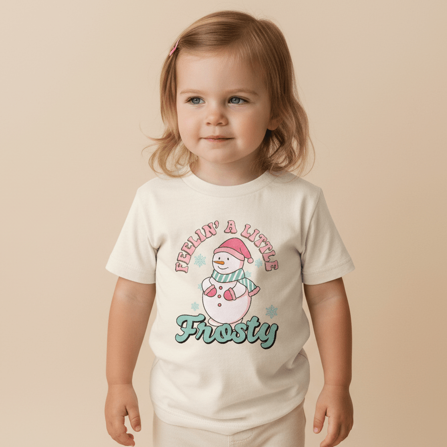 A young child with light brown hair wears the Toodleroos Feelin’ a Little Frosty Toddler Shirt – a retro snowman tee that features a cartoon snowman with a red hat and scarf, along with the phrase “Feelin’ a Little Frosty” printed above and below the image.