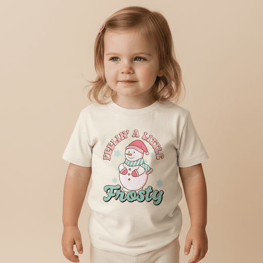 A young child with light brown hair wears the Toodleroos Feelin’ a Little Frosty Toddler Shirt – a retro snowman tee that features a cartoon snowman with a red hat and scarf, along with the phrase “Feelin’ a Little Frosty” printed above and below the image.