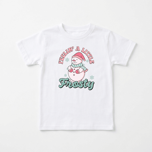 This Toodleroos “Feelin’ a Little Frosty Toddler Shirt – Retro Snowman Tee” features a white tee with a cartoon snowman wearing a Santa hat and scarf, surrounded by small snowflakes. The text “Feelin’ a Little” appears above the snowman, with “Frosty” below, making it the perfect vintage-inspired holiday shirt for toddler photos.