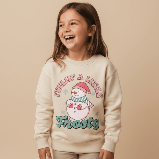 A smiling young girl wears the Toodleroos Feelin’ a Little Frosty Toddler Sweatshirt – Retro Snowman Sweatshirt, featuring a cheerful snowman graphic, playful snowflakes, and the phrase “Feelin’ A Little Frosty” on the front. The scene is set against a neutral beige background.
