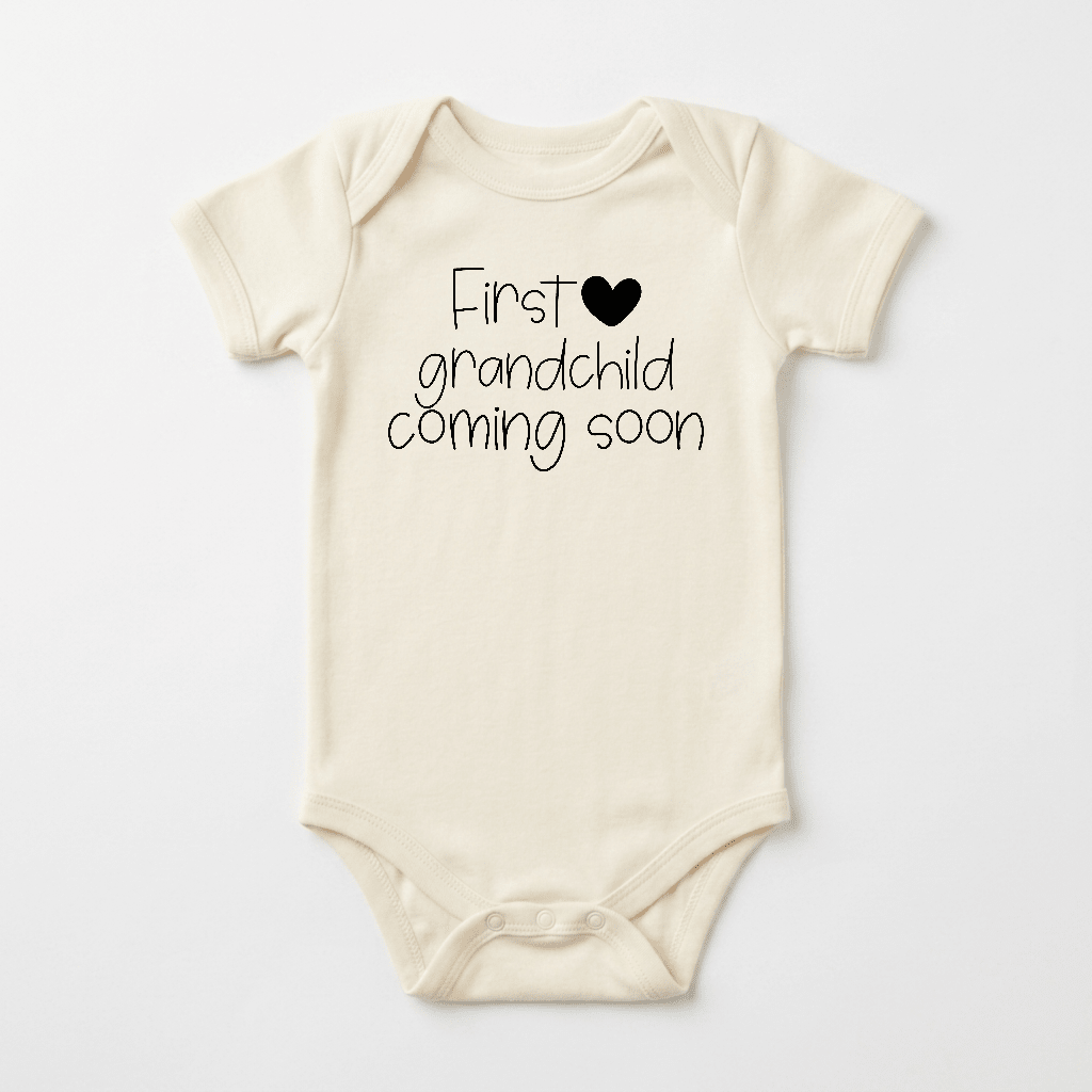 Cream-colored First Grandchild Coming Soon Baby Onesie – Grandparent Announcement by Toodleroos, featuring bold black text and a heart design, laid flat on a white background. This charming Toodleroos onesie is the perfect choice for an unforgettable pregnancy reveal to grandparents.