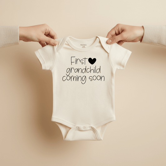Two hands hold up a white Toodleroos First Grandchild Coming Soon Baby Onesie – Grandparent Announcement, featuring a black heart on the front, making it the ideal outfit for a pregnancy reveal against a neutral beige background.