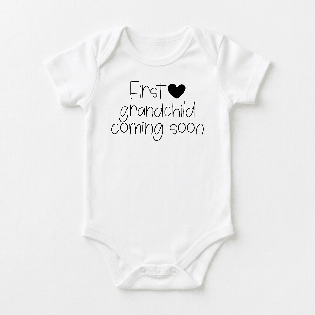 Introducing the Toodleroos First Grandchild Coming Soon Baby Onesie – a white baby bodysuit featuring black text and a heart symbol on the front, perfect for sharing your exciting news as a grandparent announcement or baby reveal outfit.