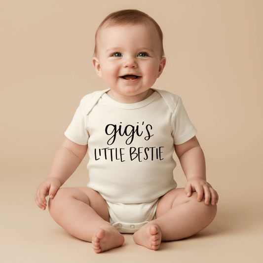 A smiling baby sits on a beige background wearing the Toodleroos Gigi’s Little Bestie Baby Onesie – Handwritten Grandma Bodysuit, featuring “gigis little bestie” in playful black handwriting—an adorable choice for honoring the grandma and baby bond or gifting at baby showers.