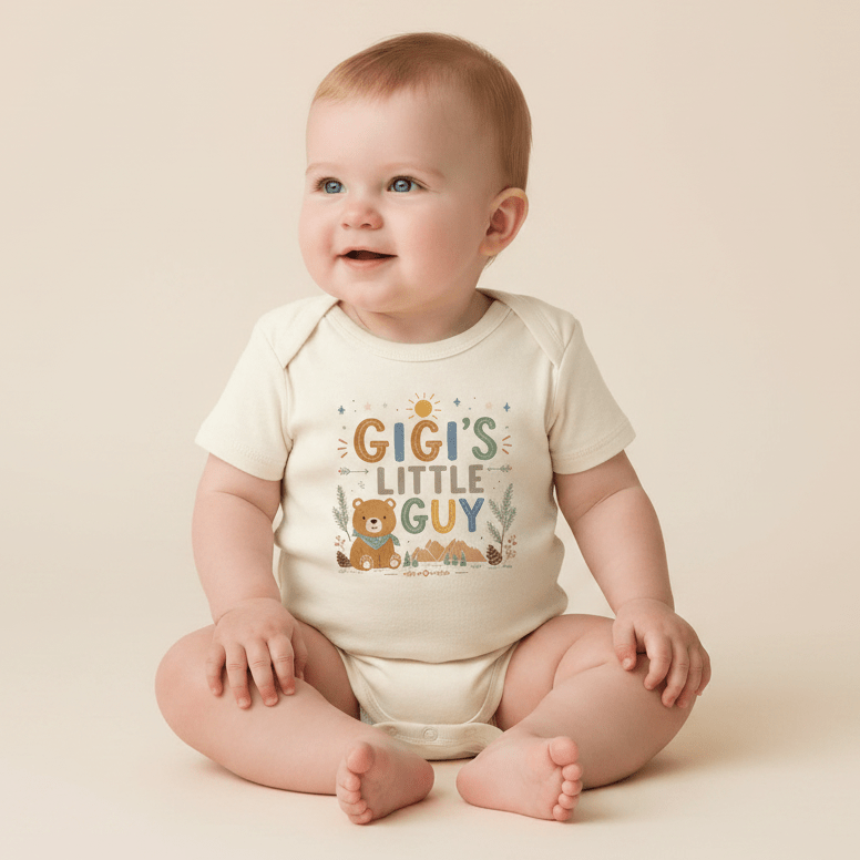 A smiling baby in a cream Toodleroos Gigi’s Little Guy Baby Onesie – Woodland Bear Design sits on the floor. The onesie showcases vibrant text reading “Gigi’s Little Guy,” adorned with playful bear, trees, sun, and mountain illustrations. This adorable bodysuit makes a thoughtful baby shower gift or a special outfit for any grandson. Neutral background.