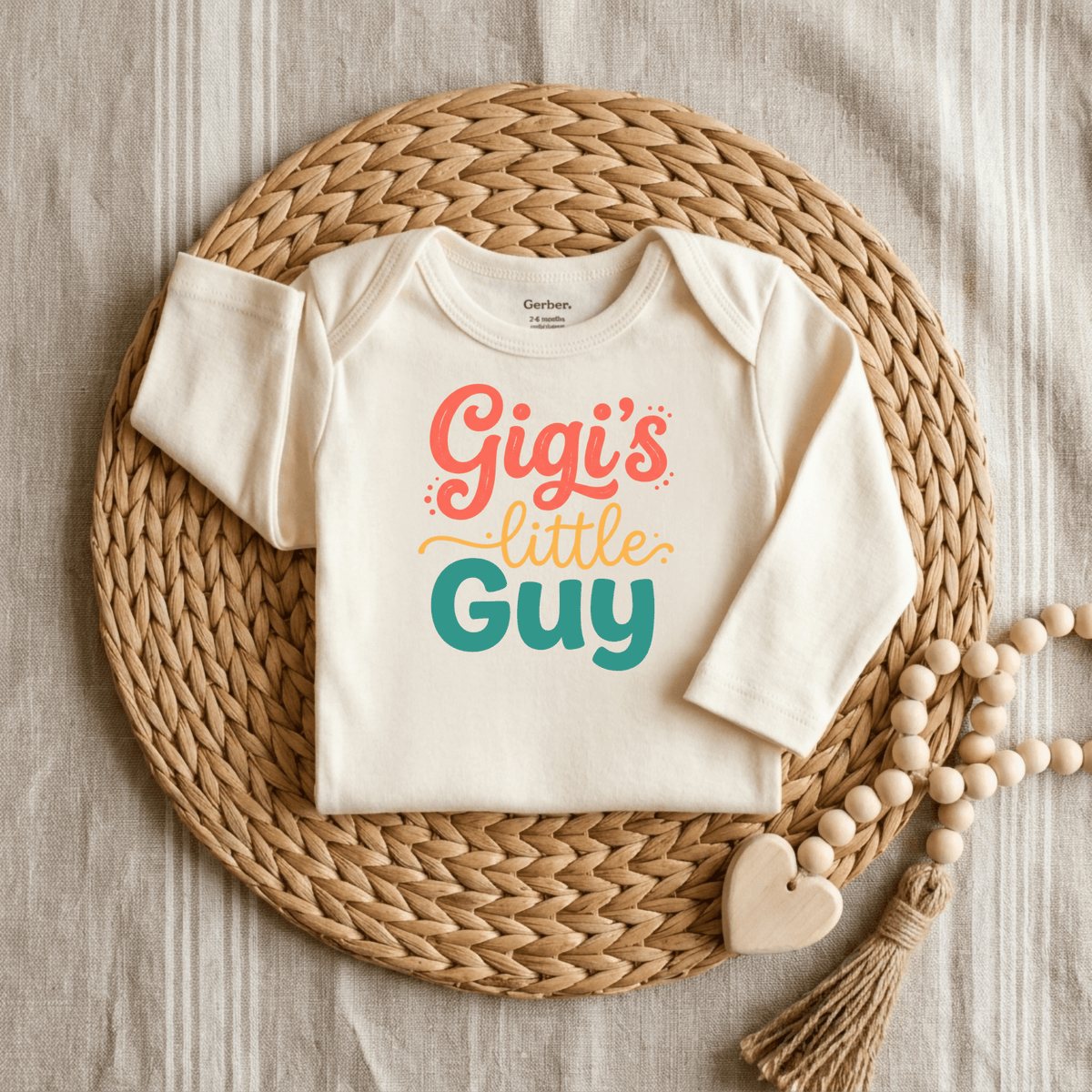 A cream-colored Gigi’s Little Guy Onesie – Baby Boy Bodysuit by Toodleroos, featuring the words “Gigi’s little guy” in colorful letters, is displayed on a woven placemat with a beaded heart decoration beside it, making it the perfect Gigi onesie and a thoughtful baby shower gift.