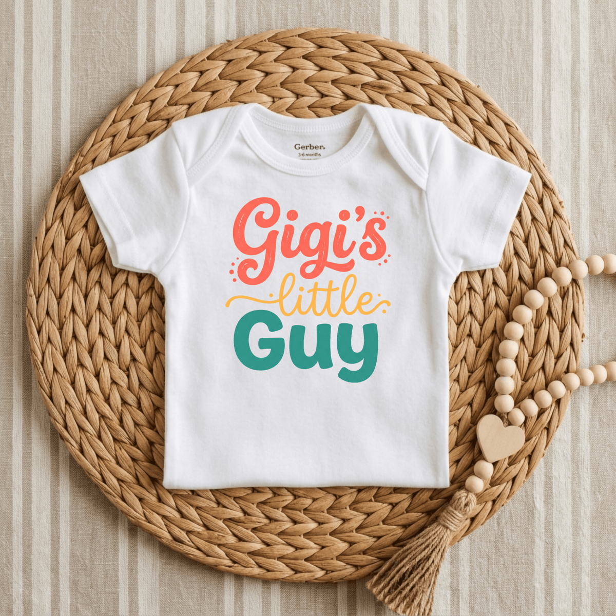 A Toodleroos “Gigi’s Little Guy Onesie – Baby Boy Bodysuit” in white, featuring colorful lettering, is displayed on a woven mat beside a beaded tassel with a wooden heart atop striped fabric—making it an adorable baby shower gift for little boys.