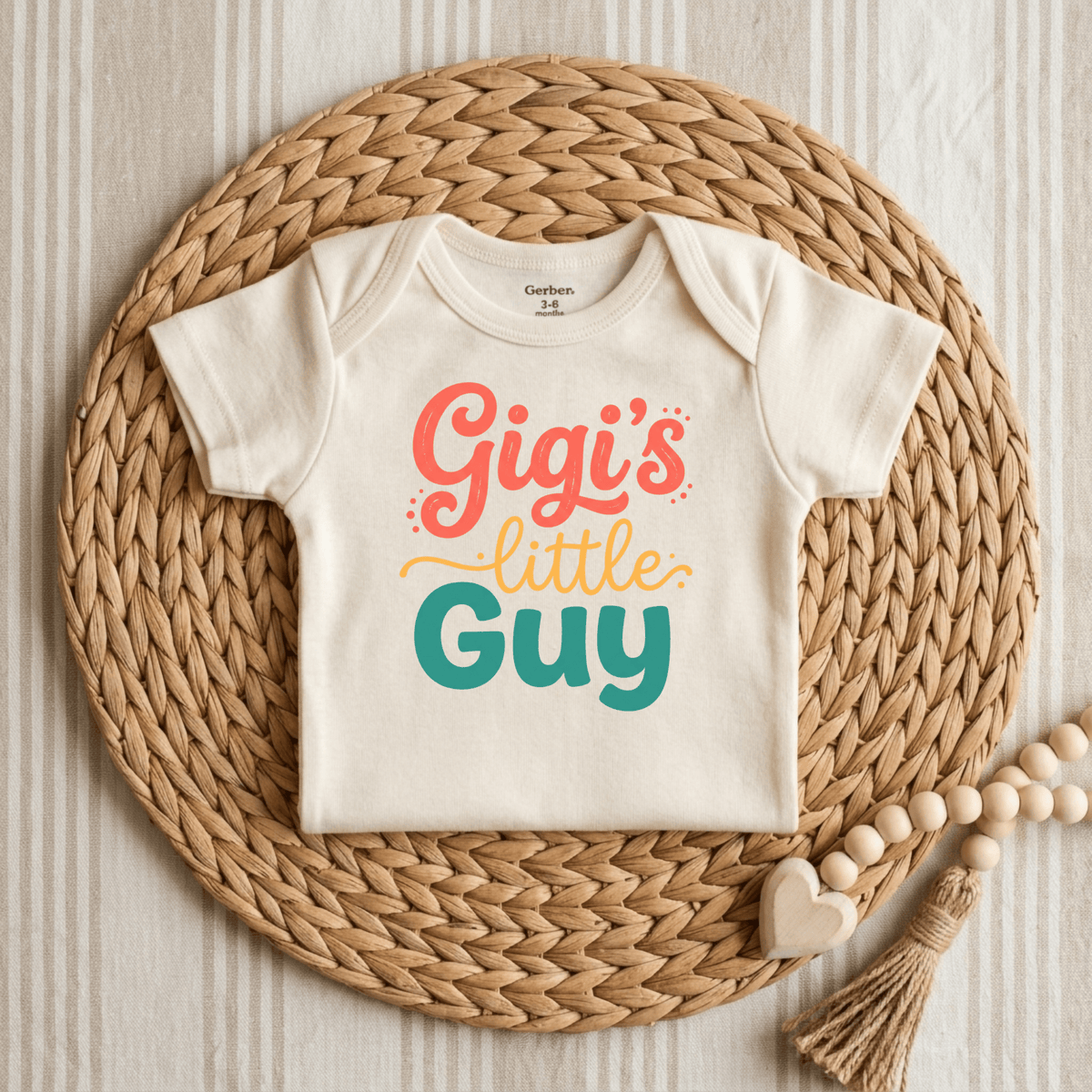 A cream Toodleroos Gigi’s Little Guy Onesie – Baby Boy Bodysuit is displayed on a woven placemat, featuring the phrase “Gigi’s little guy” written in colorful letters. Wooden beads and a heart-shaped accent sit beside it on a neutral striped surface, making this Gigi onesie an ideal baby shower gift.