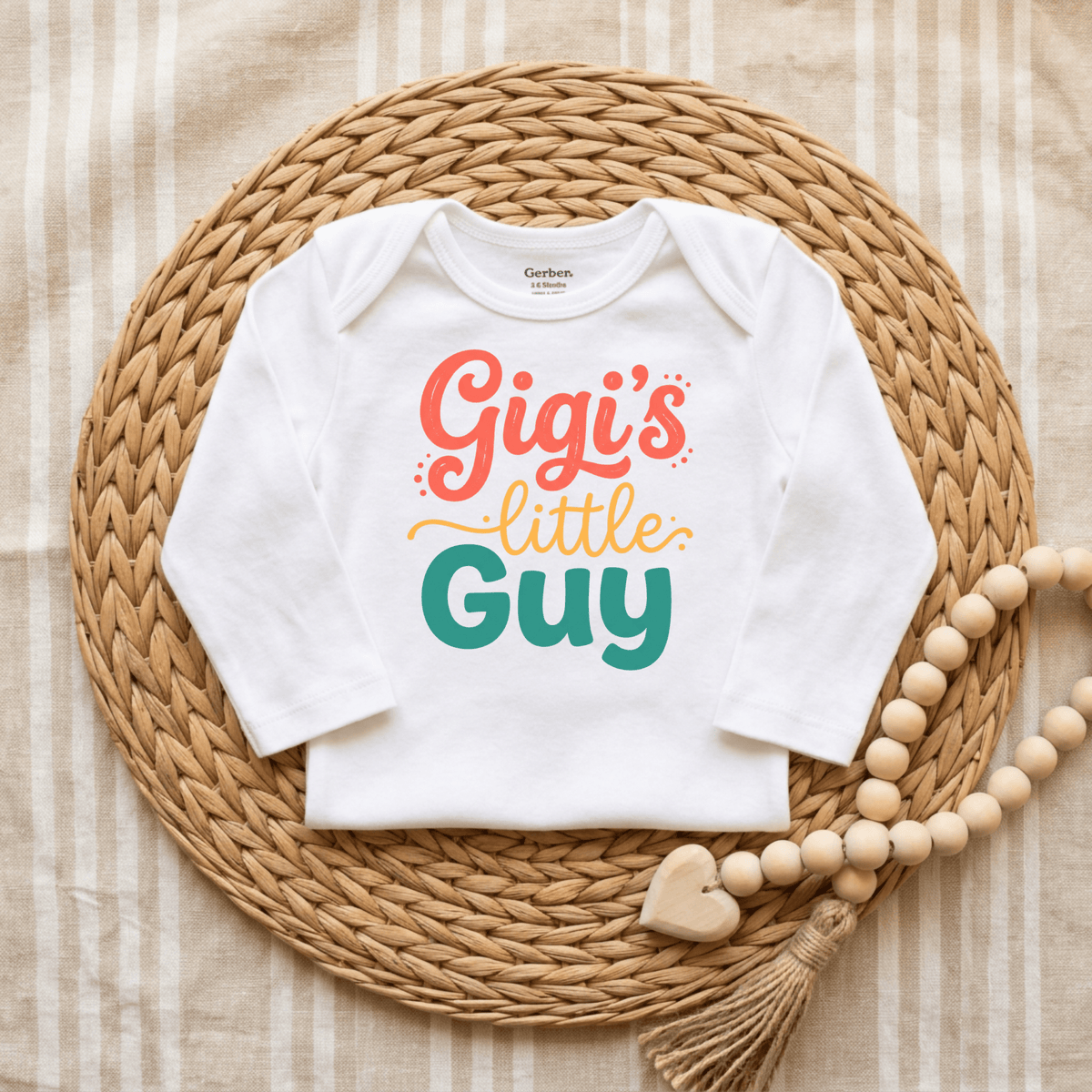 The Toodleroos Gigi’s Little Guy Onesie – Baby Boy Bodysuit features the phrase “Gigi’s little guy” in colorful, playful letters on a crisp white fabric. Displayed on a woven placemat with charming wooden bead and heart decorations, this adorable bodysuit makes a perfect baby shower gift for any little boy.