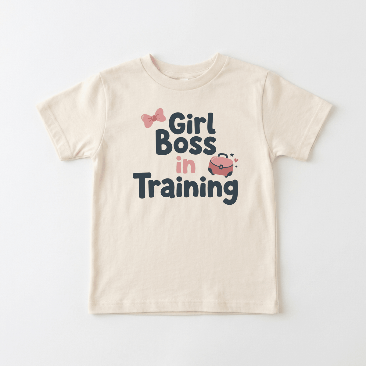 The Toodleroos “Girl Boss in Training” Toddler Girls Shirt – Confident Bow Tee is a beige graphic tee featuring bold lettering, an adorable pink bow, and a sparkling pink purse—perfect for little ones who love stylish girls’ shirts.