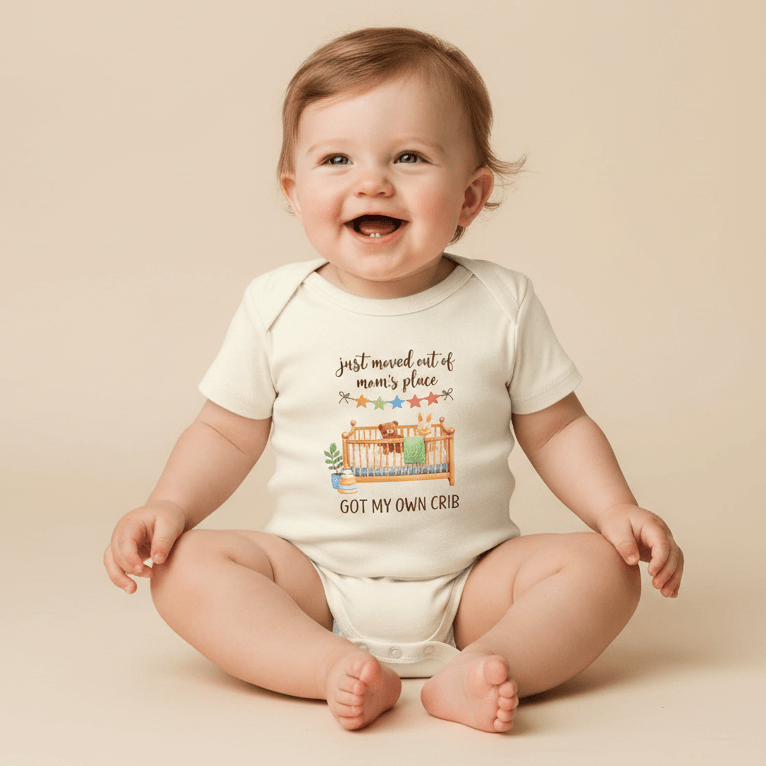 A smiling baby sits on the floor wearing the Toodleroos Funny Baby Onesie – Got My Own Crib Short-Sleeve Infant Bodysuit, featuring a playful crib illustration and the phrase “Just moved out of mama’s place, got my own crib” on the front—an adorable choice that makes a perfect baby shower gift.