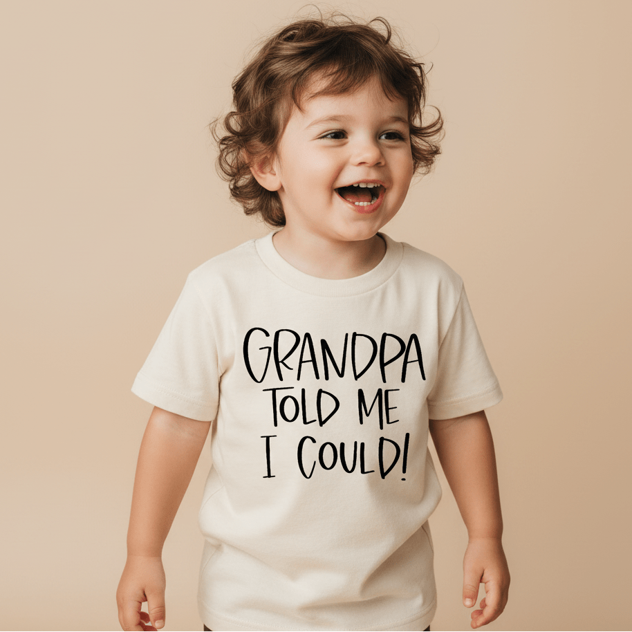 A happy toddler wears the Toodleroos “Grandpa Told Me I Could Toddler Shirt – Funny Family Tee” in cream on a neutral beige background, featuring the playful phrase, “Grandpa told me I could!”.