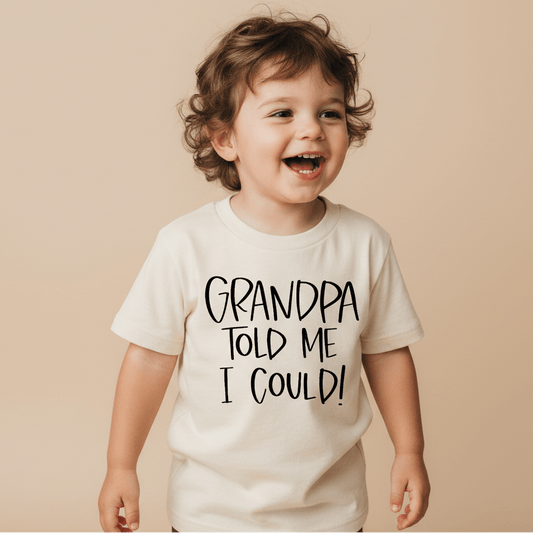 A happy toddler wears the Toodleroos “Grandpa Told Me I Could Toddler Shirt – Funny Family Tee” in cream on a neutral beige background, featuring the playful phrase, “Grandpa told me I could!”.