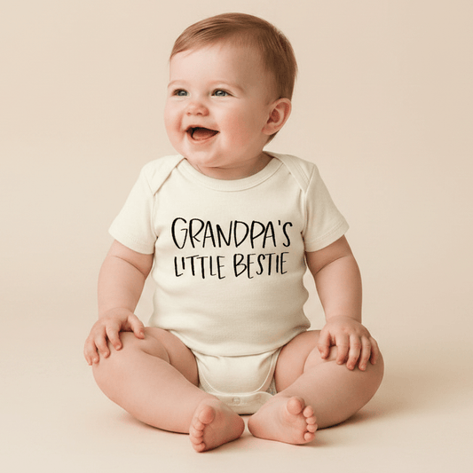 A smiling baby sits on a light-colored floor, wearing the Toodleroos Grandpa’s Little Bestie Baby Onesie – Handwritten Grandpa Bodysuit, featuring “Grandpas Little Bestie” in bold black letters—a heartwarming baby shower gift idea from Grandpa.