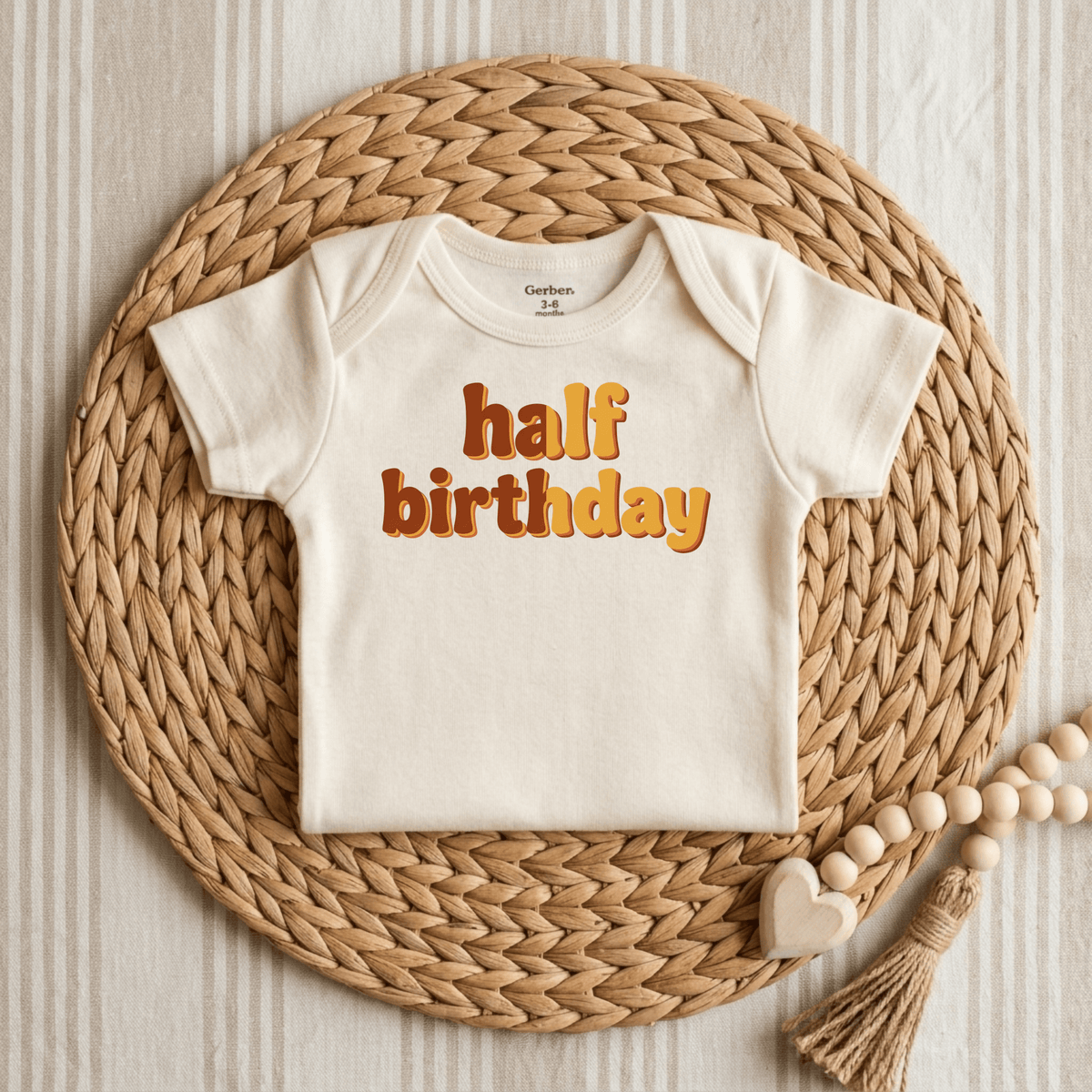 Celebrate your baby’s special day with the Toodleroos Half Birthday Onesie – Cute 6 Month Milestone Bodysuit. This retro-inspired cream onesie features “half birthday” printed in playful brown and orange letters, beautifully displayed on a woven mat with wooden beads and a heart accent, set against a striped fabric background—making it the perfect milestone photo outfit for capturing unforgettable memories.