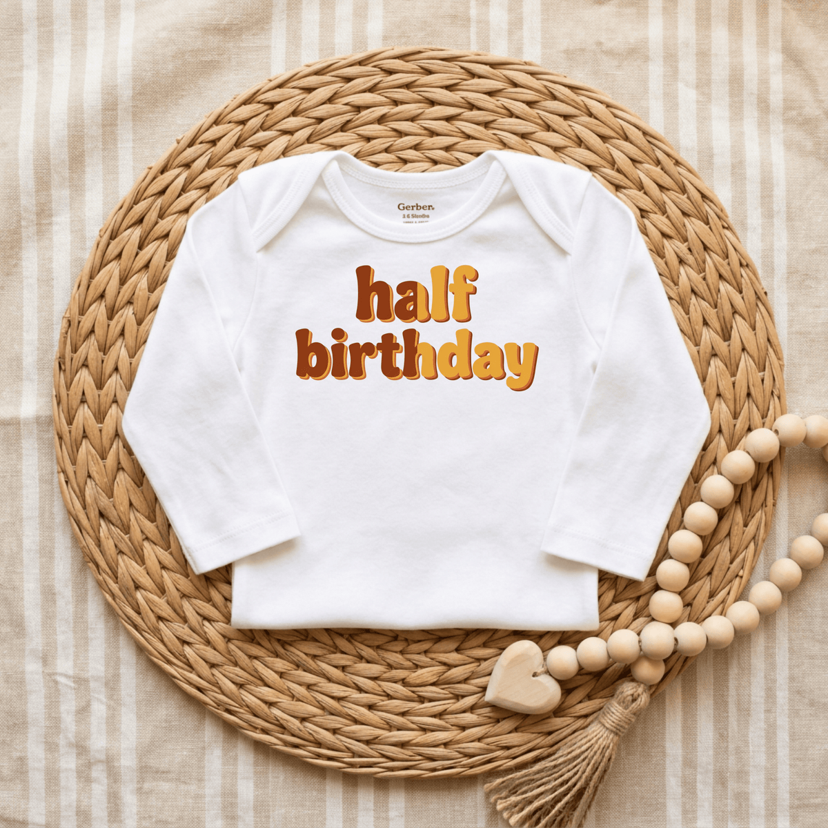 A Toodleroos “Half Birthday Onesie – Cute 6 Month Milestone Bodysuit” with vibrant orange lettering is displayed on a woven mat, complemented by a neutral beaded garland and heart charm, all set against a striped fabric backdrop—making it the perfect milestone photo outfit.
