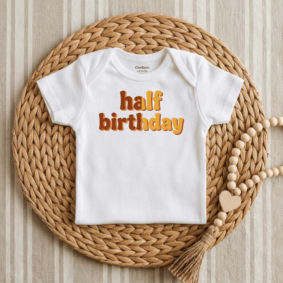The Toodleroos Half Birthday Onesie – Cute 6 Month Milestone Bodysuit is featured as a white baby bodysuit with “half birthday” in playful orange lettering, making it an adorable choice for milestone photos or a retro-themed outfit. It’s displayed on a round woven placemat beside a wood bead garland, set against a striped fabric background.
