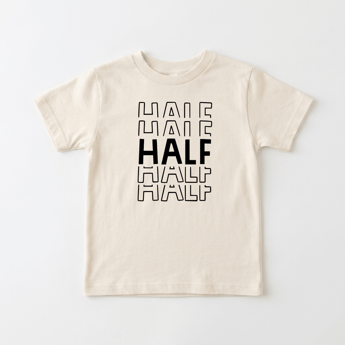 Introducing the Toodleroos Half Birthday Kids Shirt – 6 Month Milestone Tee! This beige t-shirt features the word HALF printed four times in black; the center HALF is bold, while the others are outlined for a fun contrast. Perfect as a half birthday shirt, this unique black and white kids tee makes an adorable milestone photo shirt for celebrating that special 6-month mark.