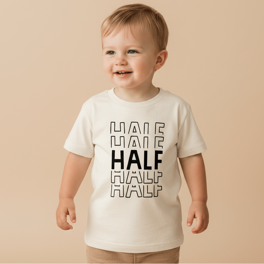 A smiling toddler stands in front of a plain beige background, wearing the Toodleroos Half Birthday Kids Shirt – 6 Month Milestone Tee. The black and white shirt features the word HALF printed four times in black, with the middle HALF bolded—making it the perfect outfit for half birthday milestone photos.