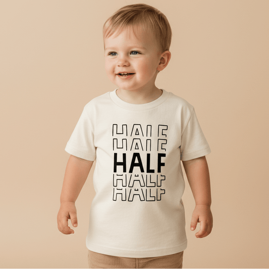 A smiling toddler stands in front of a plain beige background, wearing the Toodleroos Half Birthday Kids Shirt – 6 Month Milestone Tee. The black and white shirt features the word HALF printed four times in black, with the middle HALF bolded—making it the perfect outfit for half birthday milestone photos.