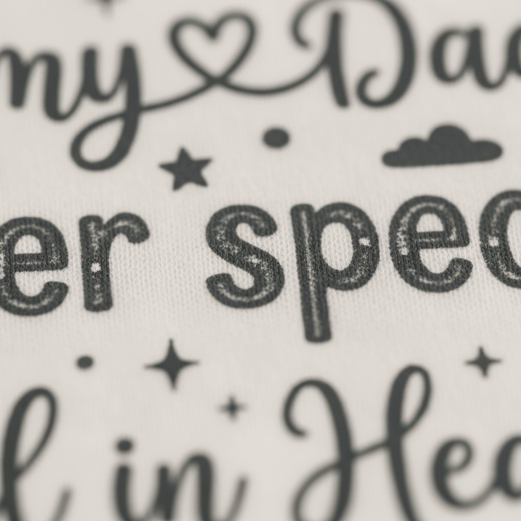 Close-up of black decorative text on white fabric, featuring the phrase “Handpicked by My Angel Grandma” with stars and a heart in the design—perfect as a memorial baby bodysuit or angel grandma onesie from Toodleroos.