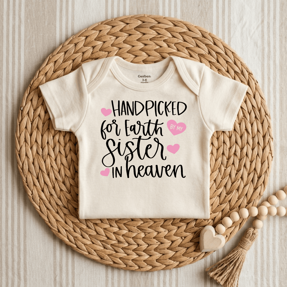 The Toodleroos Handpicked for Earth by My Sister in Heaven Baby Onesie is shown in cream, featuring black and pink text, styled on a woven placemat with a wooden bead heart garland next to it.