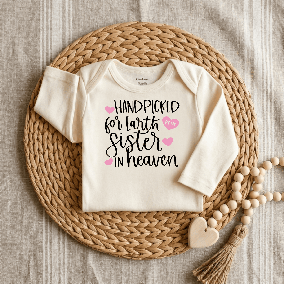 The Toodleroos Handpicked for Earth by My Sister in Heaven Baby Onesie is a cream memorial bodysuit featuring the touching phrase “Handpicked for Earth by my sister in heaven” in black and pink lettering. Displayed on a round woven mat next to a wooden bead heart garland, this sibling remembrance onesie offers a heartfelt tribute to the enduring bond between siblings, honoring your little one’s special guardian angel.