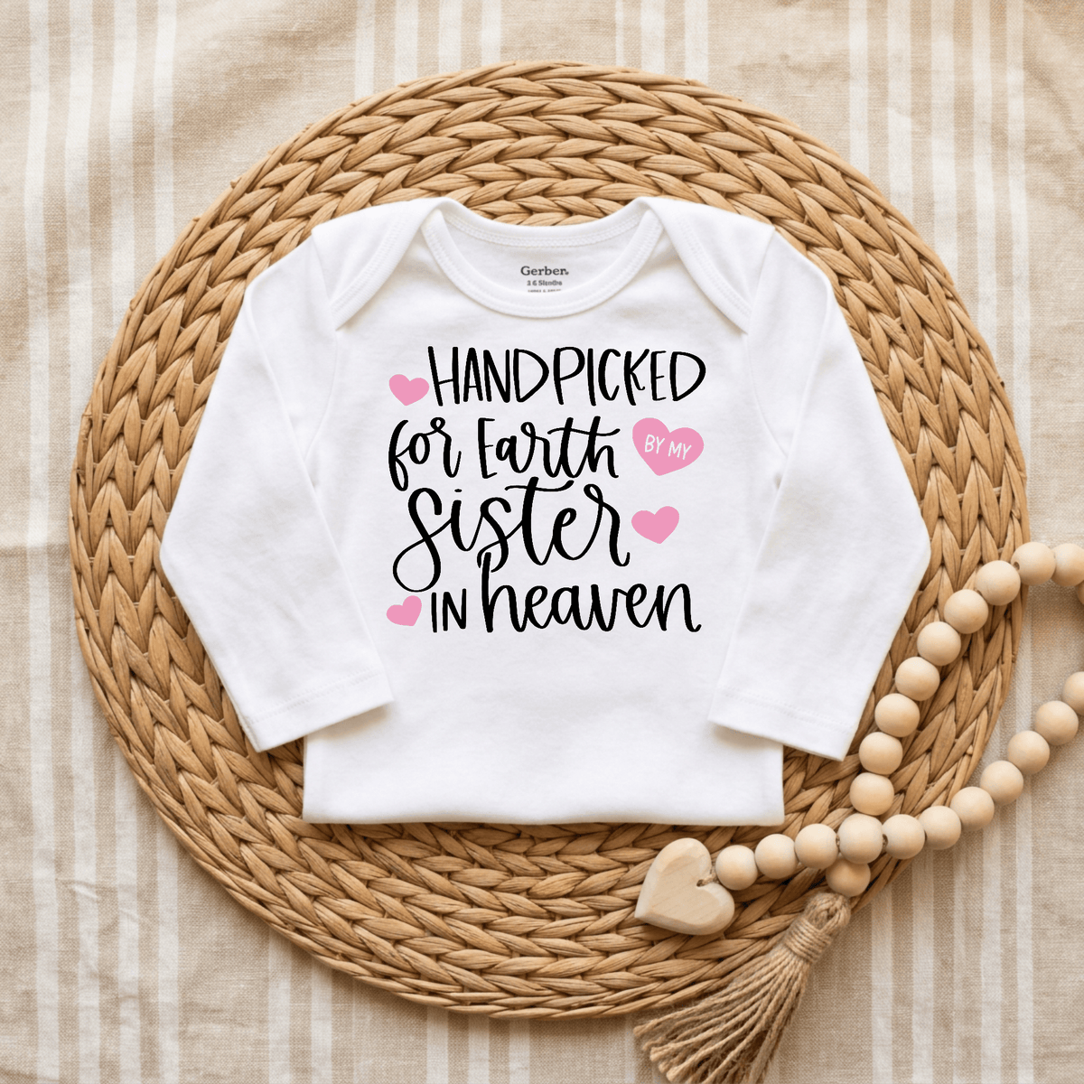A white Toodleroos Handpicked for Earth by My Sister in Heaven Baby Onesie rests on a woven mat, featuring the heartfelt message adorned with pink hearts. This sweet sibling remembrance bodysuit is accompanied by a wooden bead garland with a heart, creating a touching tribute.